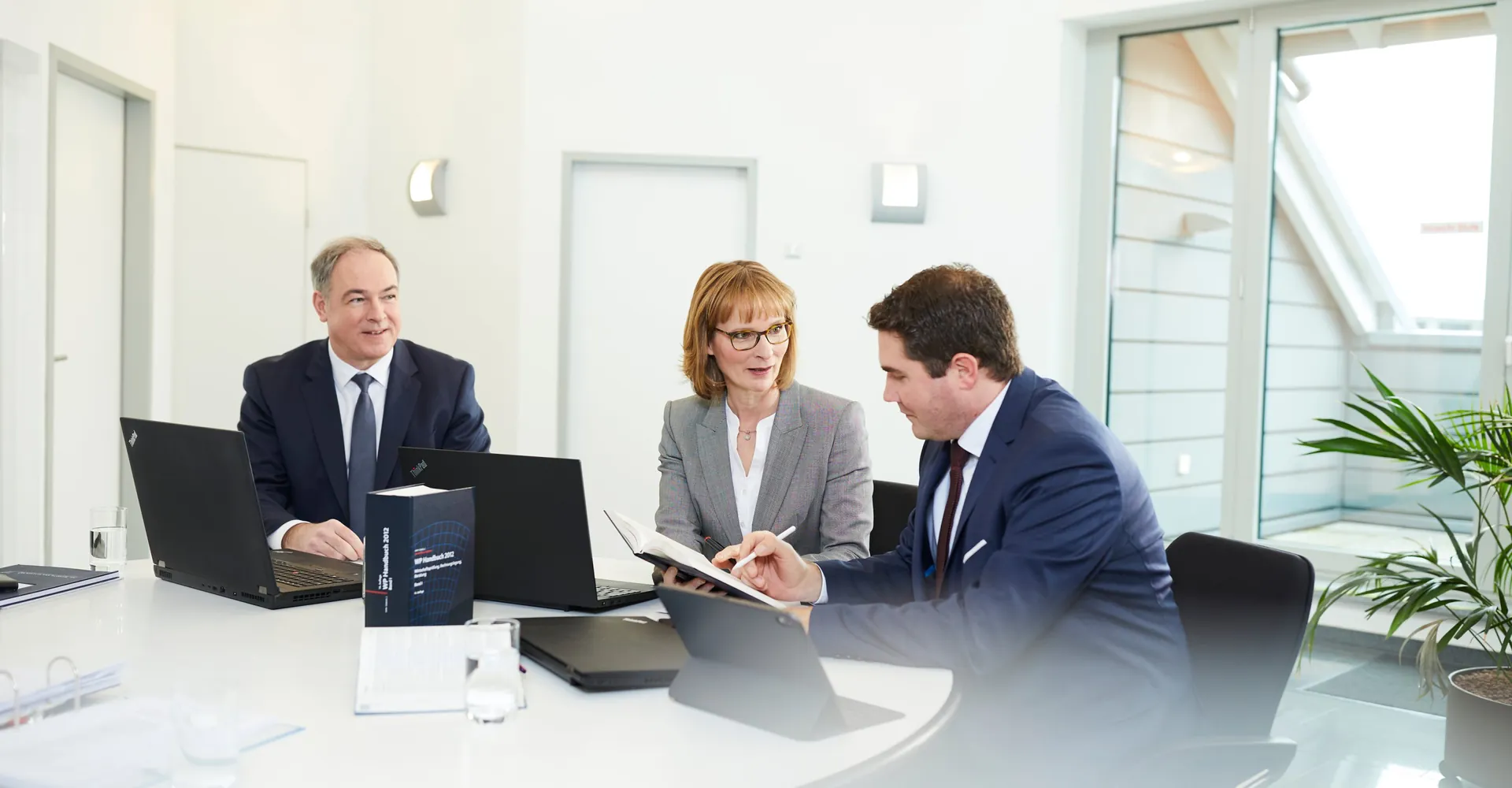 Drei Personen besprechen am runden Tisch einer hellen Kanzlei Vertragsunterlagen